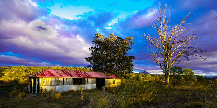 Abandoned house with a red roof in a field under a colorful sky.