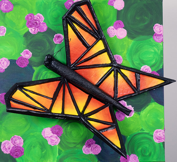 Butterfly-shaped sculpture on a canvas with orange and yellow wings on a green and purple floral background.